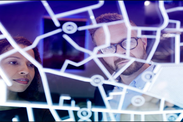 A black businesswoman and a white businessman look at a chart with dots and lines on a high tech glass screen