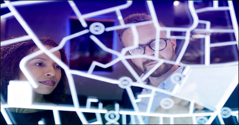 A black businesswoman and a white businessman look at a chart with dots and lines on a high tech glass screen