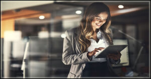 a white professional woman with lon brown hair smiles at a tablet in an office setting.