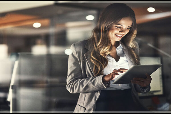 a white professional woman with lon brown hair smiles at a tablet in an office setting.