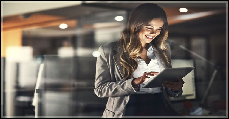 a white professional woman with lon brown hair smiles at a tablet in an office setting.