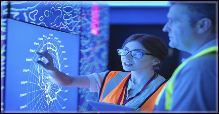 a young woman points to a readout on a digital screen of operational technology and a man watches.