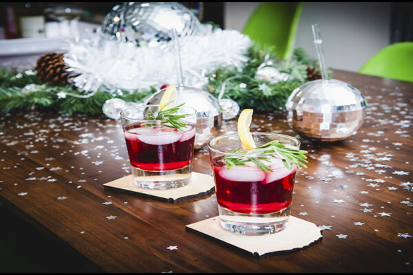 two rocks glasses half filled with red liquid sit on a dark wood table surrounded by shiny star confetti, fake snow and greenery