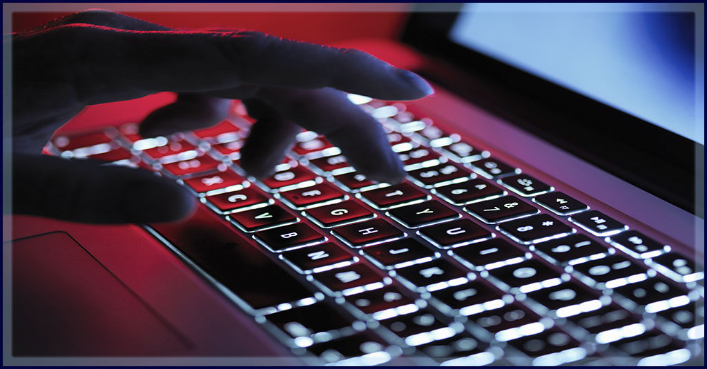 a shadowy caucasioan hand types on a backlit laptop in a dim room