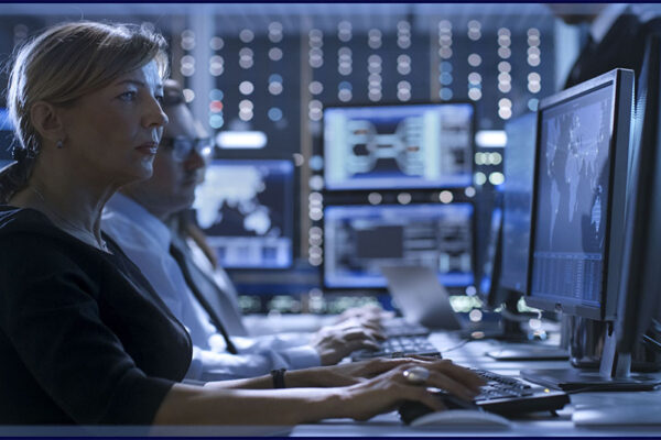 blonde Caucasian woman analyst types on a keyboard in a room full of screens of data